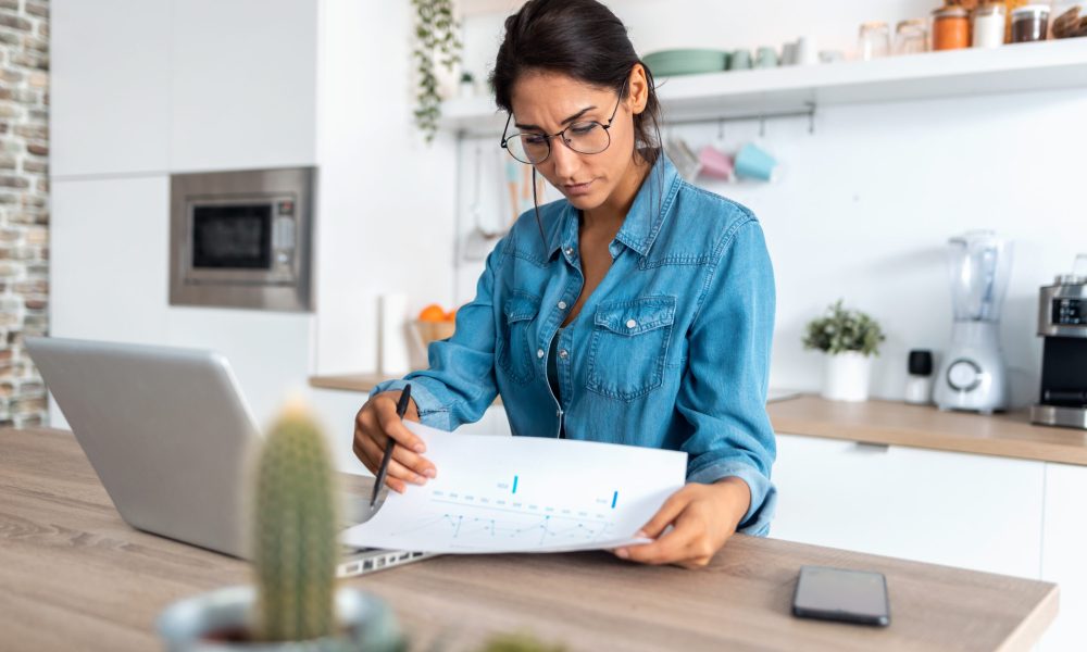 Shot of pretty young woman reviewing paperwork and working on a laptop in the kitchen at home.